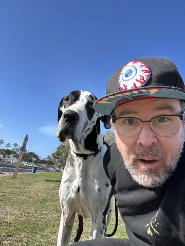Raymond in a cap and glasses beside a harlequin Great Dane outdoors, Long Beach dog walker