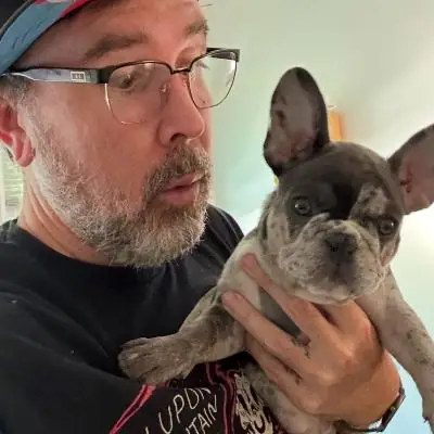 Raymond in a cap and glasses holding a fawn French bulldog indoors, Long Beach dog walker